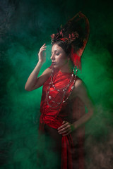strict and scary girl in a fashionable long red dress and a voluminous hat on the background of green smoke in the Studio at night alone. Halloween concept.