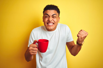 Young brazilian man drinking cup of coffee standing over isolated yellow background screaming proud and celebrating victory and success very excited, cheering emotion