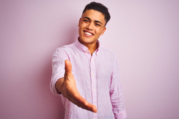 Young brazilian man wearing shirt standing over isolated pink background smiling friendly offering...