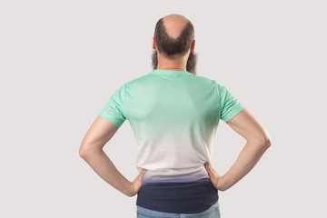 backside portrait of middle aged bald man with beard in light green t-shirt standing with hands on waists. indoor studio shot, isolated on grey background.