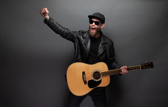 Singer and guitar player singing on a stage. He is a rocker and he is wearing leather biker jacket or asymmetric zip jacket with black cap, jeans.