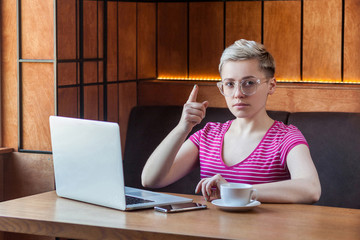 Portrait of serious young adult woman with blonde short hair, in pink shirt and eyeglasses is...
