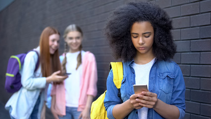 Upset black student smartphone, laughing classmates standing behind, bullying - obrazy, fototapety, plakaty