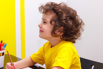 Little curly boy in yellow t-shirt sits at a table, smiles, has a rest after the letter, eyes are closed.