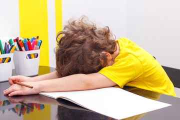Tired little boy sitting at table, with hishead on his arm, writing in her notebook while doing homework