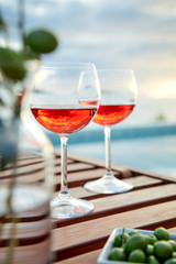 two glasses of rose wine with  olives against blue water on a pier..