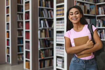 Cheerful afro girl student standing next to bookshelves