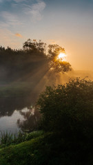Warm autumn sunrise with sun rays at the river with trees and mist at the foggy morning