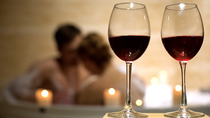 Red wine glasses standing on background of beloved couple enjoying in bathtub