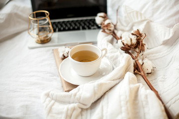 A white mug with tea, cotton, a laptop, a plaid, a candle and a book stand on the bed. Breakfast in bed. Cozy. Autumn. Winter.