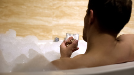 Man resting and relaxing in comfortable bathtub drinking whiskey with ice, spa