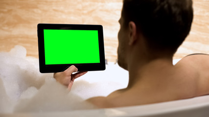 Guy reading news on green-screen tablet resting in bathtub at home, relax