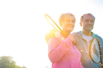 Active senior couple holding badminton racket in park with yellow lens flare in background
