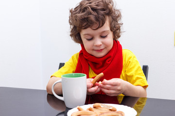 Little curly boy drinking from cup of hot drink tea in cold weather, sitting at home in scarf in kitchen at the table