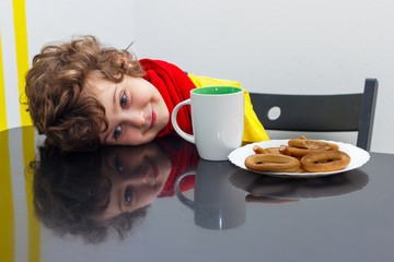 Little curly boy drinking from cup of hot drink tea in cold weather, sitting at home in scarf in kitchen at the table