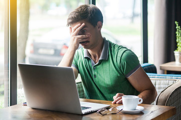 Spy, shy or afraid! Young businessman in green t-shirt sitting try to looking at laptop display through his fingers. business and freelancing concept. indoor shot near big window at daytime.
