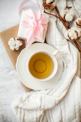 White mug with tea, cotton, plaid, book and white gift box with a pink ribbon on the bed. Breakfast in bed. Cozy. Autumn. Winter.