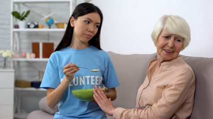 Upset elderly woman refusing to eat oatmeal offered by volunteer, nursing home
