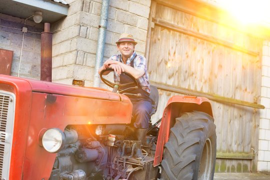 Smiling mature farmer driving tractor in barn with yellow lens flare in background