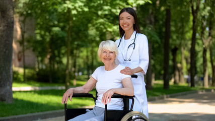 Female nurse and disabled aged woman smiling to camera, relax in hospital park