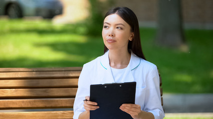 Fototapeta premium Thoughtful female doctor working outside, sitting on bench in hospital park