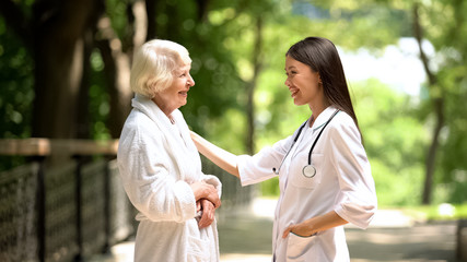 Smiling nurse talking with elderly woman in bathrobe at sanatoria park, relax - obrazy, fototapety, plakaty