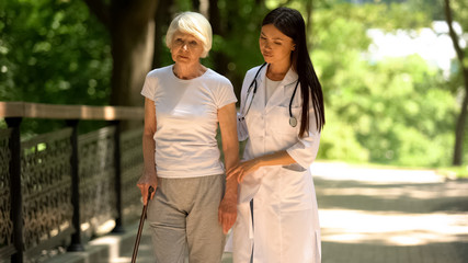 Doctor and upset old female patient with cane walking in hospital park, health