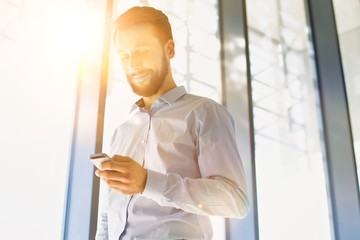Portrait of young businessman using smartphone in office hall with yellow lens flare in background