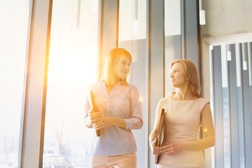 Businesswomen discussing plans while walking in office hall with yellow lens flare in background
