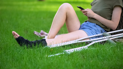 Woman in ankle brace sitting park with crutches scrolling smartphone, fracture