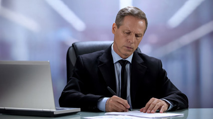 Attentive company boss reading and signing documents at office table with laptop