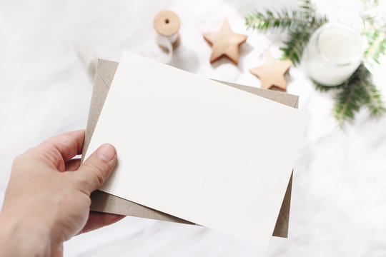 Closeup of woman's hand holding blank paper card. Greeting card mock-up scene. Chritsmas winter design. Feminine styled stock photo. Blurred background with fir branch, candle and wooden stars.