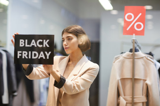 Waist up portrait of beautiful woman hanging Black Friday sale sign on window display in clothes store, copy space