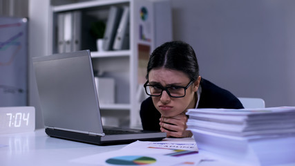 Upset female office employee looking desperately at pile of paper lying on desk