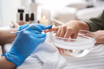 Young woman soaking hand in nail bath while manicurist using cuticle pusher - obrazy, fototapety, plakaty