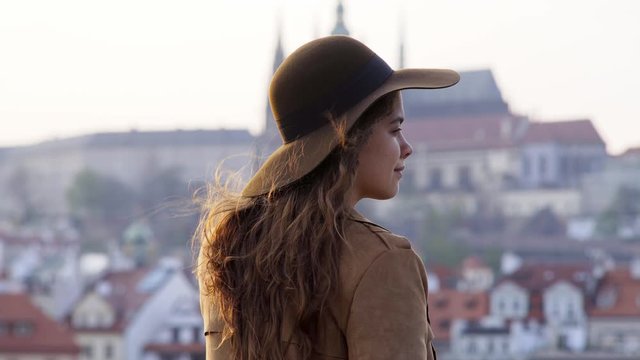 Back view female tourist enjoying beautiful view historical centre of Prague. Attractive girl in hat standing in backlit and looking at Prague Castle on defocused background