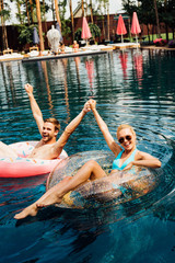 happy couple holding hands while lying on swim rings in swimming pool