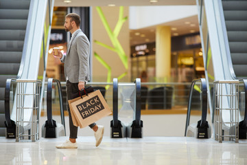 Side view portrait of fashionable man holding shopping bags with Black Friday inscription walking across hall in mall, copy space