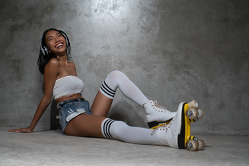 Young beautiful Asian girl in retro quads roller skates posing in studio over concrete wall...