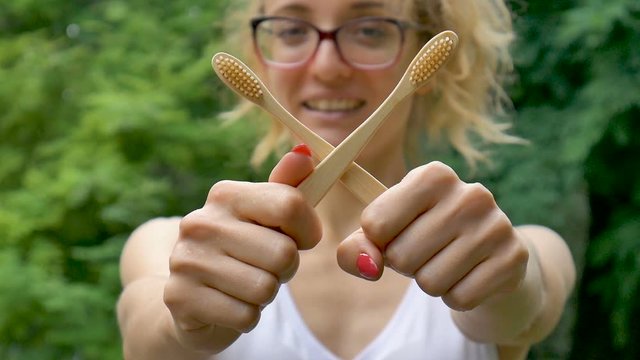 Young beautiful girl with ponytail is holding a useful bamboo toothbrushes outdoors during rainy weather on green tree background. Environmental friendliness and zero waste concept.