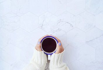 Top view of woman hands holding hot cup of coffee on white marble background, warm sweater in winter concept