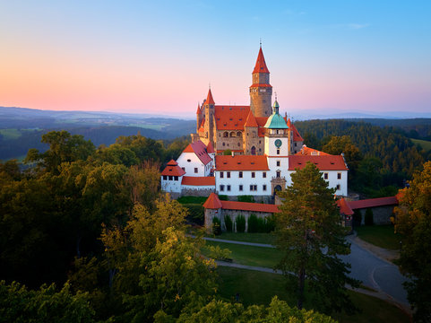 Aerial view on a romantic fairytale castle on a knoll in highland landscape, surrounded by trees. Gothic castle with high towers, red roofs, stone walls. Bouzov castle, Moravian region, Czechia.