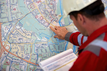 Energy industry. A technician dressed in red overalls and a white helmet stands in front of a city map. Checking the parameters of the urban heating system. Start of the heating season.
