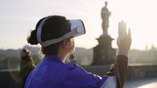 Close-up portrait woman 80's style clothes standing in backlit on street. Girl wearing VR headset, turning head around , touching virtual objects at defocused background old european town
