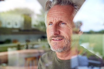 Senior Man Standing And Looking Out Of Kitchen Door Viewed Through Window