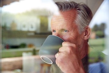 Senior Man Standing And Looking Out Of Kitchen Door Drinking Coffee Viewed Through Window