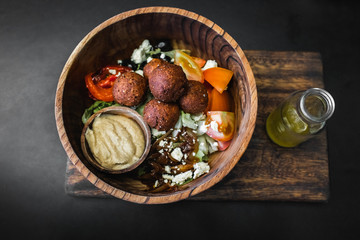 Fototapeta na wymiar Wooden bowl with falafel balls, hummus and fresh salad. Olive oil in bottle on side. Dark black background, top view. Traditional israeli food. Veganism concept.