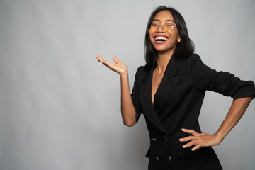 Portrait of attractive young Asian smiling woman in elegant black blazer and glasses pointing hand aside over light gray wall background. Mock up copy space - obrazy, fototapety, plakaty