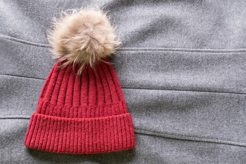 Cozy and warm winter flat lay with copy space. Dark red knitted hat with fur pompom on gray woolen crumpled background