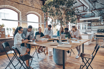 Group of young modern people in smart casual wear communicating and using modern technologies while working in the office
